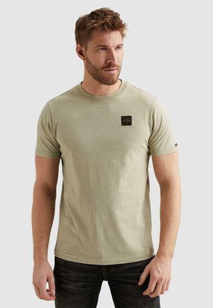 Man with beard wearing a light olive green short-sleeve t-shirt with a small black patch on the left chest and dark jeans, looking right.
