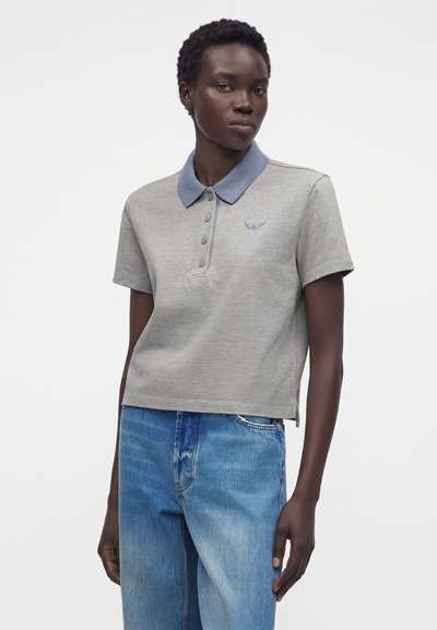 Γυναίκα με σκουρόχρωμο δέρμα που φοράει ανοιχτογκρι πόλο μπλούζα με μπλε γιακά και μπλε έμβλημα, συνδυασμένη με μπλε ψηλόμεσα τζιν, στέκεται μπροστά σε λευκό φόντο.