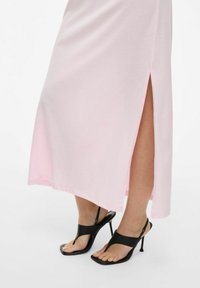 Jasnoróżowa, sięgająca do kolan sukienka z bocznym rozcięciem, wykonana z miękkiego materiału, połączona z czarnymi sandałami na wysokim obcasie z odkrytymi palcami w kwadratowym kształcie.