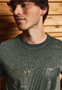 Tmavě zelené tričko s texturovaným povrchem, zdobené grafickým potiskem, který obsahuje slovo "sníh" a diagonální pruhy v antracitově šedé.