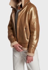 Guldmetaljacka med en fluffig beige krage och manschetter, med en ficka med dragkedja och slät lädertextur. Vita byxor kompletterar looken.