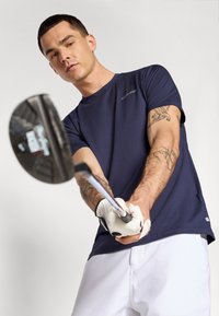 Sötétkék sportpóló rövid ujjal, Calvin Klein logóval. A modellen golfütő van, fehér golfkesztyűt és rövidnadrágot visel.