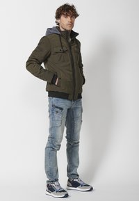 Giacca bomber trapuntata verde oliva con cappuccio grigio, chiusura con zip, due tasche sul petto, polsini a coste. Indossata con jeans cargo azzurri e sneakers.