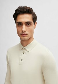Beige long-sleeve polo shirt featuring a classic collar, three buttons, and intricate stitching detail on the sleeves. Smooth texture.