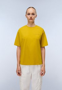 Femme avec les cheveux plaqués en arrière, portant un t-shirt jaune moutarde à manches courtes et un pantalon cargo blanc, se tenant contre un fond clair.