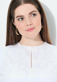 Abito bianco con un corpo in pizzo testurizzato decorato con motivi floreali, una piccola scollatura e una gonna in tessuto morbido. Orecchini a cerchio dorati completano l'outfit.