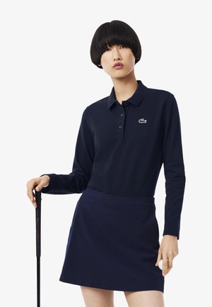 Dunkelblaues Langarm-Polo mit Kragen und grünem Logo, kombiniert mit einem passenden Rock. Der Stoff wirkt glatt und weich, ideal für Golf.