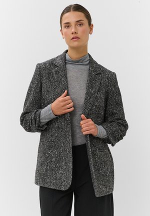 Femme portant un blazer chevrons gris sur un col roulé gris clair et un pantalon noir, debout devant un fond clair uni.