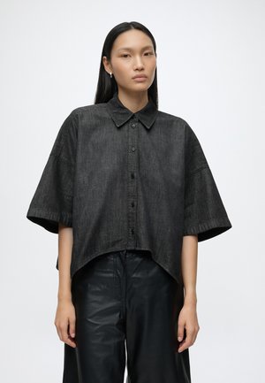 Mujer con cabello largo y negro, vistiendo una camisa negra suelta de manga corta con botones y pantalones negros, de pie contra un fondo liso.