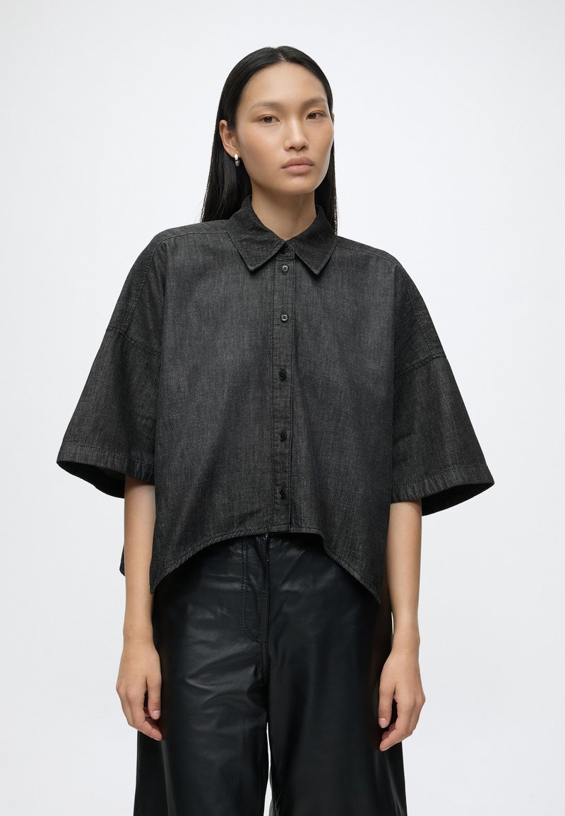 Femme aux longs cheveux noirs, portant une chemise noire ample à manches courtes avec boutons et un pantalon noir, debout devant un fond uni.