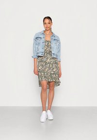 Mulher vestindo um vestido floral combinado com uma jaqueta de ganga azul clara e ténis brancos, de pé contra um fundo claro e liso.