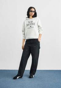 Γκρι φούτερ με κουκούλα και το γράφημα «NEW BALANCE ATHLETICS», συνδυασμένο με μαύρο παντελόνι φαρδύ με τσέπες, φορεμένο με μαύρα αθλητικά παπούτσια.