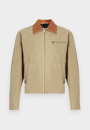 Beige zip-up jacket with brown collar, long sleeves, two side pockets, and a small zip pocket with gold medallion charm on the chest.