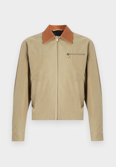 Smėlio spalvos striukė su užtrauktuku, rudu apykakliu, ilgomis rankovėmis, dviem šoninėmis kišenėmis ir maža užtraukiama kišene su auksiniu medaliono pakabuku ant krūtinės.