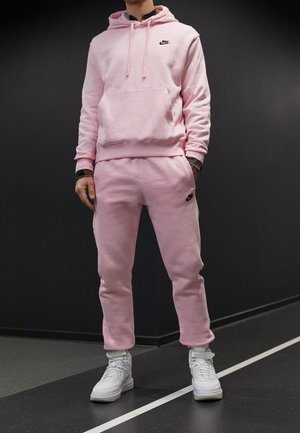 Mann trägt rosa Nike-Hoodie und passende Jogginghosen mit weißen High-Top-Sneakern, steht vor einer dunklen Wand und einem dunklen Boden.
