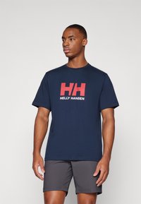 Tamno plava majica s kratkim rukavima s crvenim i bijelim "HH" logotipom sprijeda, napravljena od mekanog pamučnog materijala. Uparena s sivim šortsevima.