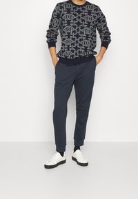 Maglione blu con un pattern geometrico grigio, abbinato a pantaloni joggers blu e sneakers bianche con suola nera.