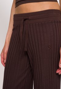Bruine geribbelde wijde broek met een drawstring taille en geborduurd logo nabij de heup. De stof heeft een zachte, gestructureerde afwerking.