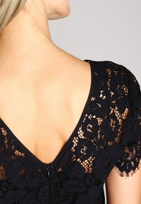 Robe en dentelle noire présentant un décolleté en V profond, des broderies florales et des bords de manches festonnés, avec une fermeture éclair discrète au dos.