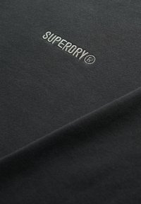 Czarna bawełniana tkanina z wyszywanym białym logo "SUPERDRY". Gładka tekstura z delikatnym połyskiem, złożona tak, aby pokazać detale wzoru.