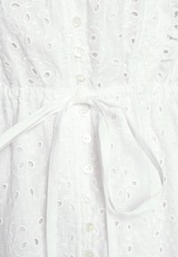 Vitt klänning med broderade pärlhålsmönster, knappstängningar och knytig midja. Texturen är lätt och med en mjuk tygfinish.