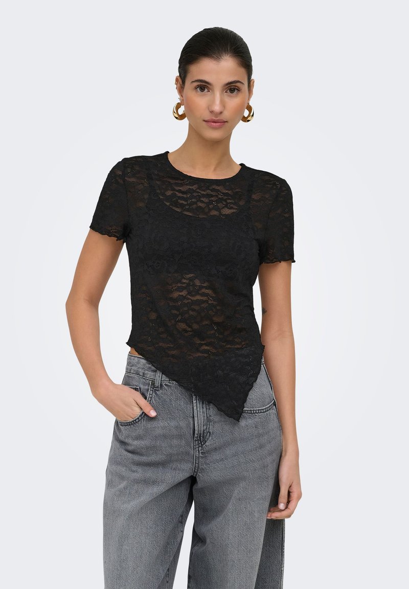 Femme aux cheveux foncés en chignon, portant des boucles d'oreilles créoles dorées, un haut en dentelle noire transparente avec un ourlet asymétrique, et un jean gris taille haute, une main dans la poche.