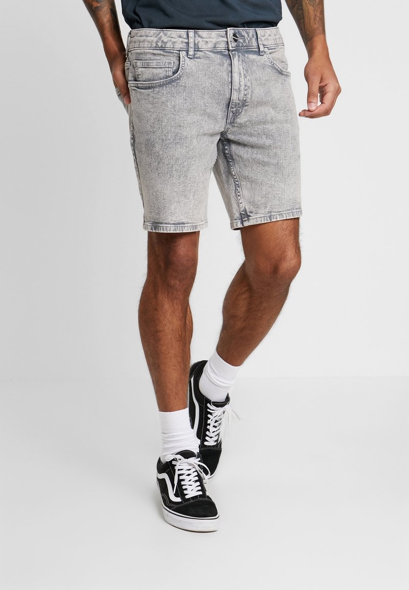 Pantalones cortos de mezclilla gris claro con un diseño clásico de cinco bolsillos. Presentan un corte recto, dobladillo vuelto y detalles de desgastado sutil. Se llevan con zapatillas negras.