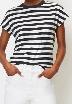 Persona con una camiseta de manga corta a rayas horizontales en blanco y negro y pantalones blancos con las manos en los bolsillos.