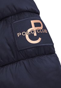 Zbliżenie na rękaw granatowej pikowanej kurtki z prostokątną naszywką z logo Polo Club w beżowych literach.