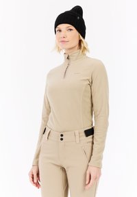 Kvinde iført beige fleece pullover og bukser med sort beanie, stående mod hvid baggrund, med ansigtet lidt til venstre.