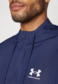 Marineblå hættejakke lavet af letvægtsmateriale, med en frontlynlås, justerbare snore og et hvidt Under Armour-logo på brystet.