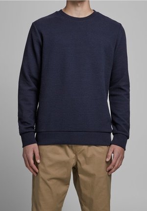 Sweatshirt bleu marine en tissu texturé, col rond, avec des poignets et un ourlet côtelés, associé à un pantalon beige en khaki.