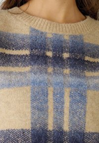 Pull en laine beige avec un motif à carreaux bleus aux nuances variées, texture douce et encolure côtelée.