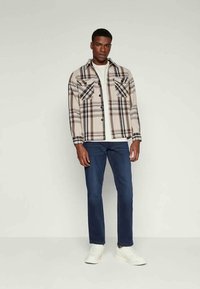 Πλαϊνός καρώ πουκάμισο σε μπεζ, καφέ και μαύρο, με δύο τσέπες στο στήθος. Συνδυασμένο με σκούρα μπλε τζιν και λευκά αθλητικά παπούτσια. Απλός σχεδιασμός.