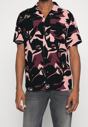 Mężczyzna noszący koszulę z krótkim rękawem zapinaną na guziki z dużym kwiatowym wzorem w kolorach różowym, czarnym i bordowym oraz szare dżinsy.