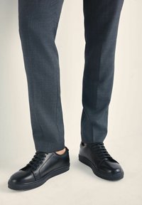 Czarne skórzane sneakersy o gładkiej fakturze, okrągłym nosku i płaskiej podeszwie, zestawione z szarymi spodniami w kant. Minimalistyczny design z sznurowadłami.