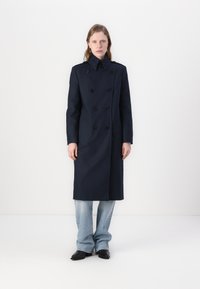 Palton marin cu două rânduri de nasturi, cu nasturi negri proeminenți, guler structurat și blugi evazați din denim deschis la culoare. Textură fină a materialului.