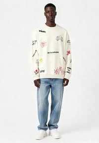 Sudadera color blanco roto con varios prints gráficos, incluyendo flores, corazones y texto. Combinada con jeans de color azul claro y zapatillas blancas.