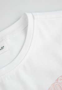 Biała bawełniana koszulka z okrągłym dekoltem. Posiada miękką teksturę oraz wyblakły różowy nadruk na przodzie, w pobliżu dolnej krawędzi.