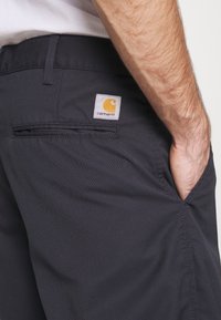 Mörkgrå arbetsbyxor med en texturerad tygkonstruktion, med en bakficka och en framträdande orange Carhartt-logotyp på midjebandet.