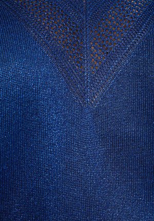 Close-up af blåt strikket stof med et V-formet netmønster i øverste midte, der viser teksturerede og perforerede strikkedetaljer.