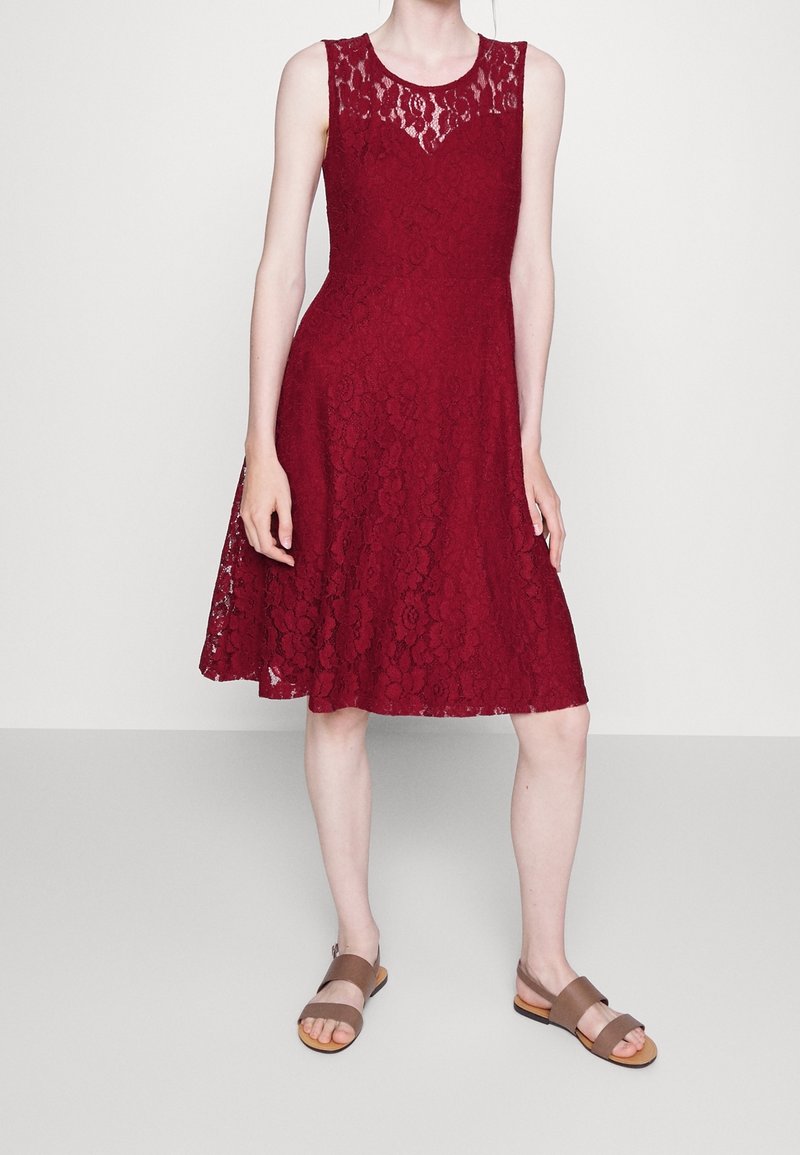 Burgundy spetsklänning med ärmlös design, som har ett texturerat mönster och en utsvängd kjol. Matchad med bruna platta sandaler.