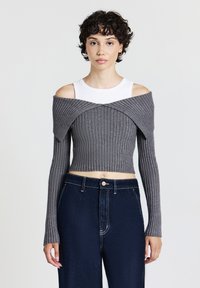 Junge Person mit kurzen lockigen Haaren trägt ein weißes ärmelloses Top unter einem grauen schulterfreien, kurzen Rippensweater und dunkelblauen Jeans.