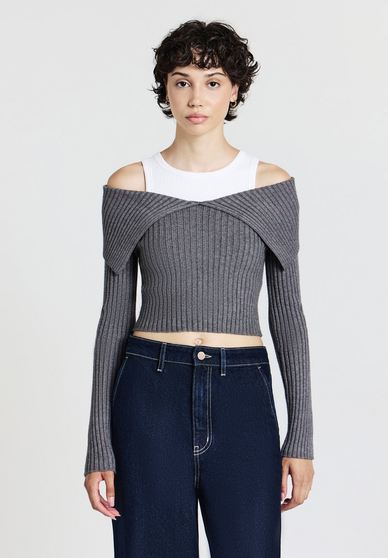 Junge Person mit kurzen lockigen Haaren trägt ein weißes ärmelloses Top unter einem grauen schulterfreien, kurzen Rippensweater und dunkelblauen Jeans.