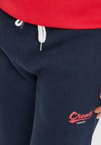 Pantalones de chándal navy con cintura elástica y cordón blanco. Presenta un logo rojo "Creek's Original" en el muslo izquierdo. Material de algodón suave.