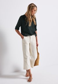 Donna che indossa un top nero a maniche lunghe, pantaloni bianchi a gamba larga e croppati, sandali beige con cinturini, tiene in mano una borsa in suede marrone, in piedi contro uno sfondo bianco.