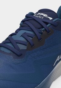 Κοντινό πλάνο ενός μπλε αθλητικού παπουτσιού με σκούρα μπλε κορδόνια, ύφασμα από δίχτυ, και ορατό λευκό λογότυπο στο πλάι.