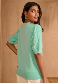 Gaiši zaļš pūkains tops ar īsām pūstām mežģīņu piedurknēm, apaļu dekoltē un rievojumu apakšmalu. Uz pleca ir dekoratīvi pogas.