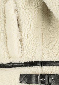Cremfarbener, strukturiert fleece Stoff mit einem schwarzen Leder Gürtel und einer Schnalle, die um die Taille angebracht ist und einen hochwertigen Materialkontrast schafft.