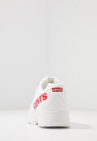 White sneaker with contrasting red "Levi's" branding. Smooth synthetic material, padded collar, and textured sole for traction.